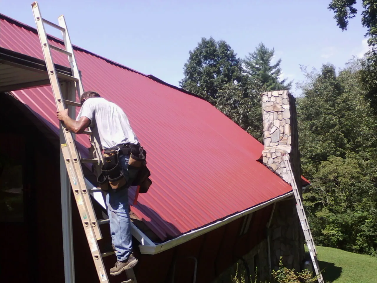 Roofer on a ladder beside a red metal roof and stone chimney — Soffit & Fascia Repair by Roofing Brothers in Netcong